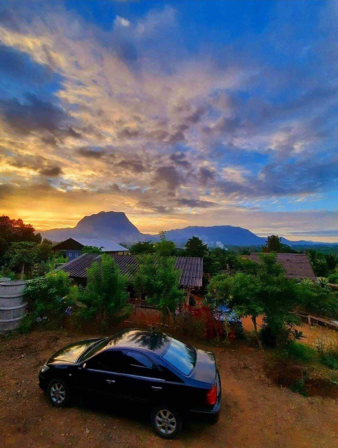 A rural landscape with a mountain in the background, a car parked in the foreground, and traditional houses with thatched roofs.