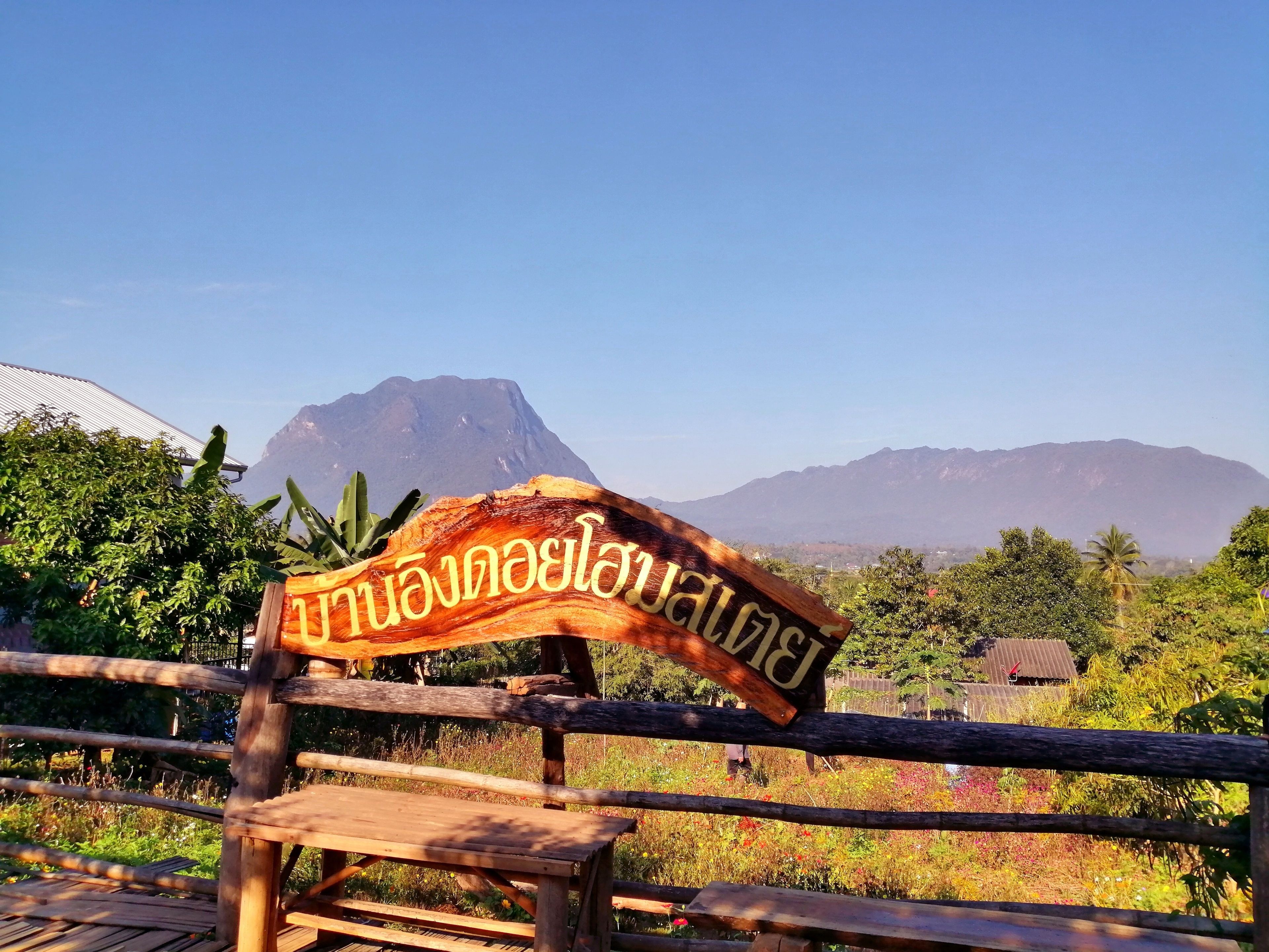 A wooden sign with Thai script, a wooden fence, and a mountainous landscape in the background.