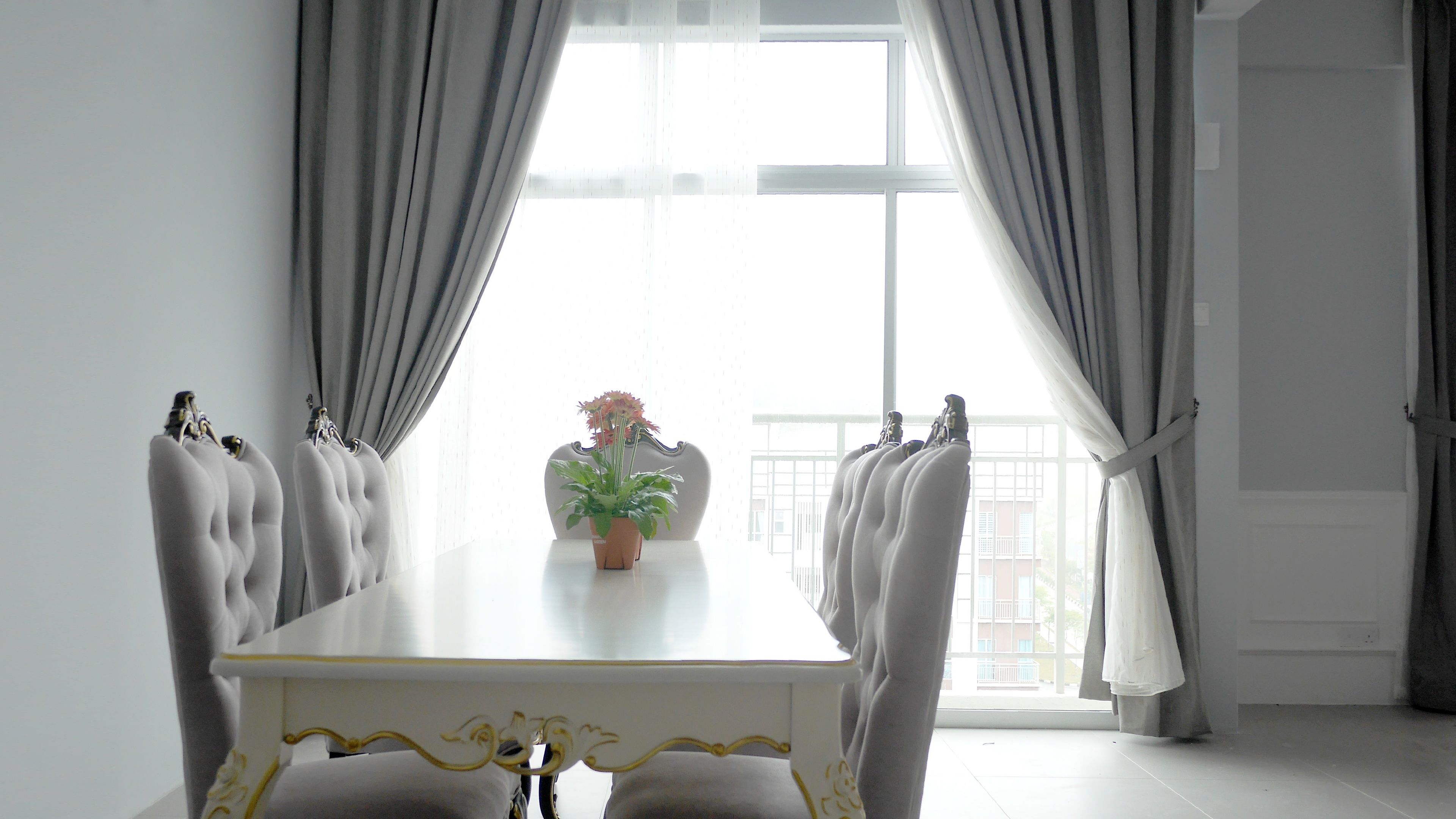 A dining area with a white table and chairs, a potted plant, and a view of a balcony through large windows.