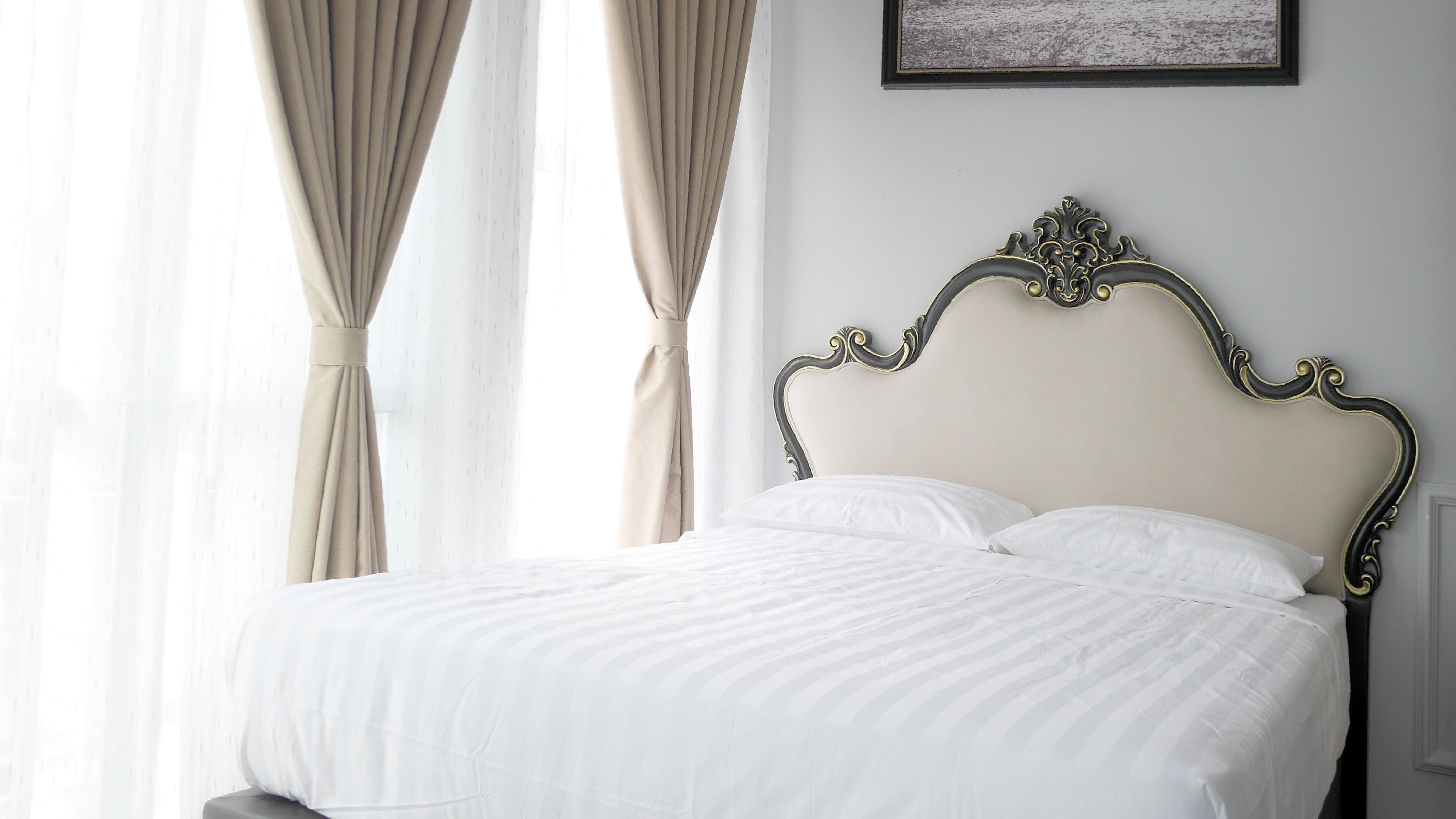 A neatly made bed with a classic headboard, a window with sheer curtains, and a framed picture on the wall.