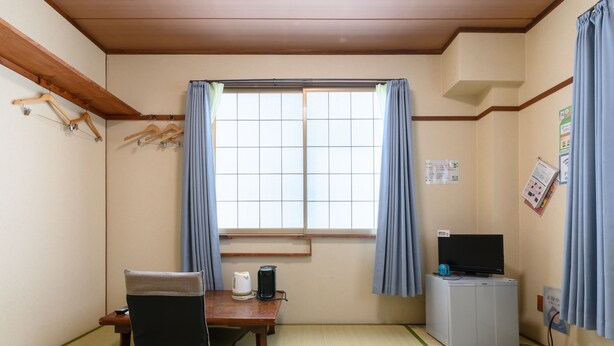Een kamer met tatami vloerbedekking, een lage tafel met een stoel, een televisie, een waterkoker en een kast.