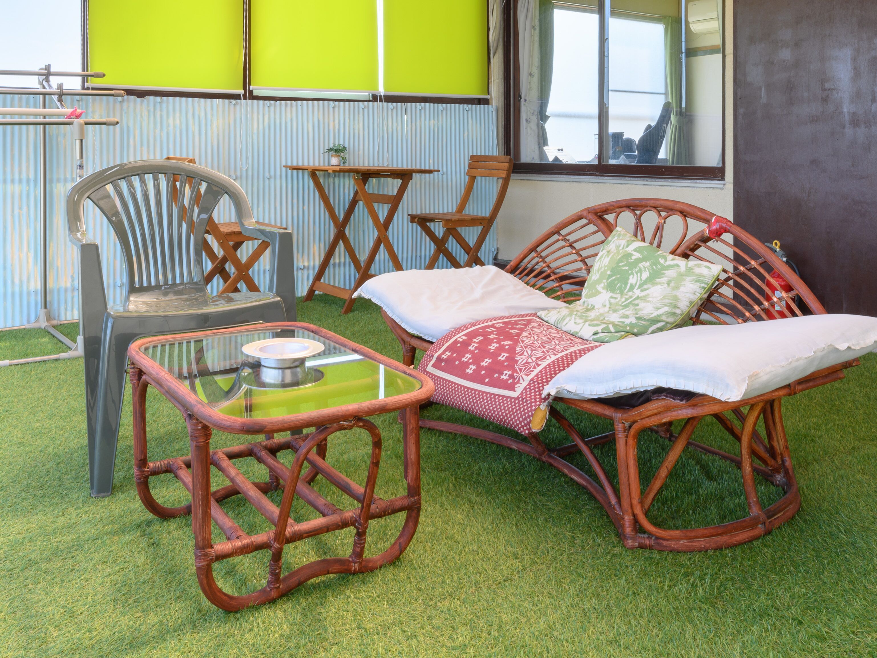 A room with a wicker sofa, a glass coffee table, and a wooden chair. There is a small table with a potted plant. The room has a green artificial grass floor and a window with curtains.