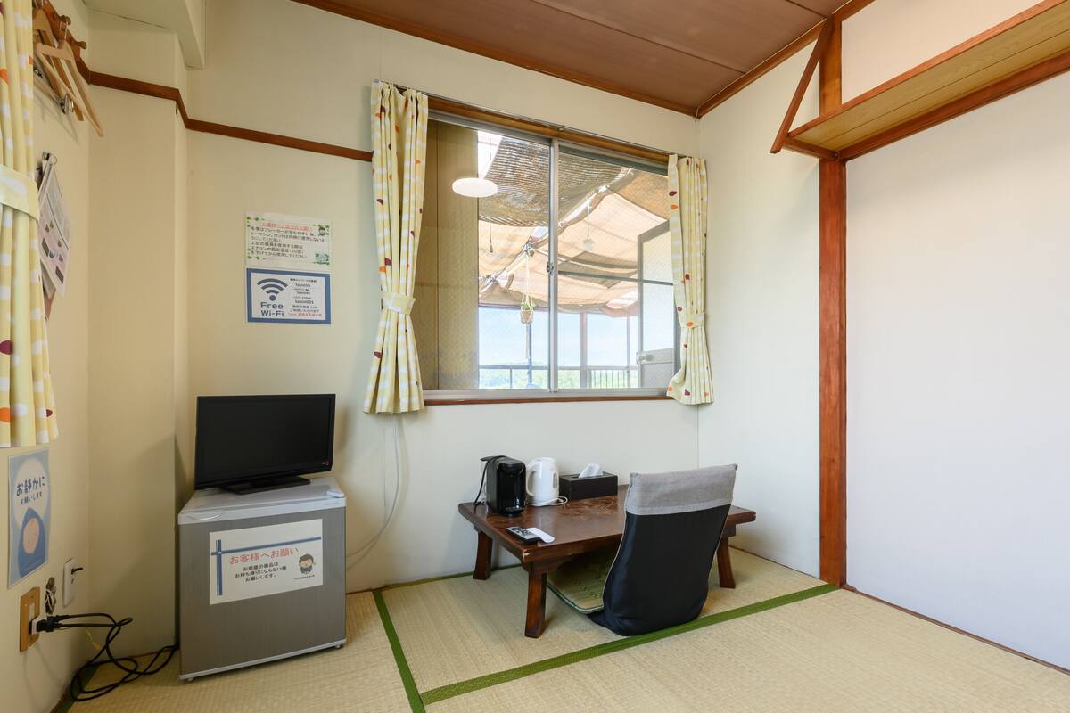 Quarto com televisão, escrivaninha com chaleira, cadeira e janela com cortinas.