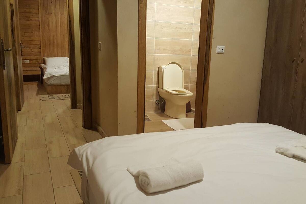 Habitación de hotel con piso de madera, cama con ropa blanca, puerta de madera y baño visible con inodoro y lavamanos.