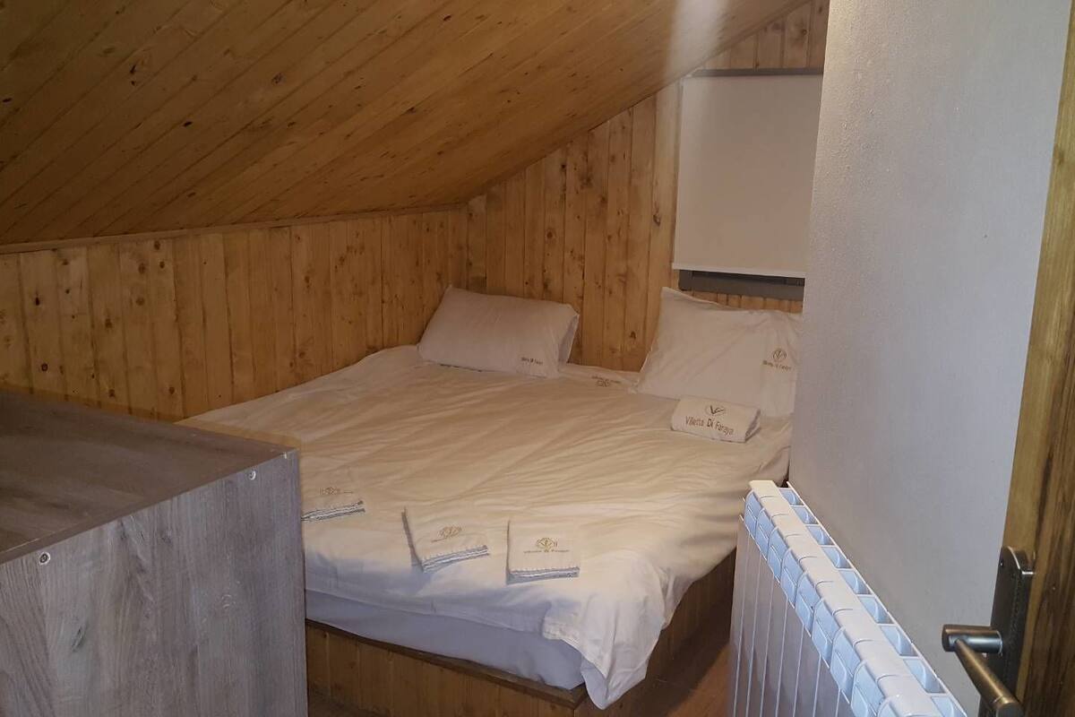 Una cama individual en una habitación de madera con colchón y almohadas blancas, una mesita de noche pequeña y un radiador.
