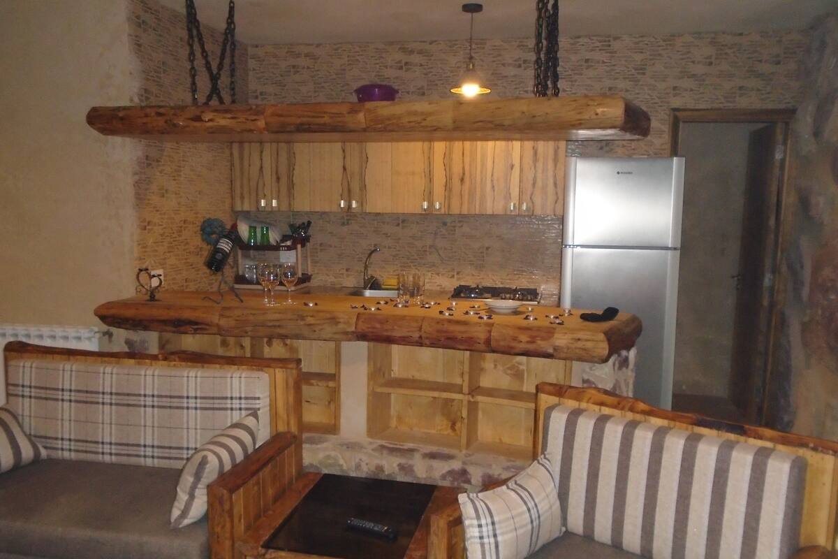 Una cocina rústica con armarios de madera, una barra empotrada y un refrigerador. Hay dos sillas de madera con cojines frente a la barra.