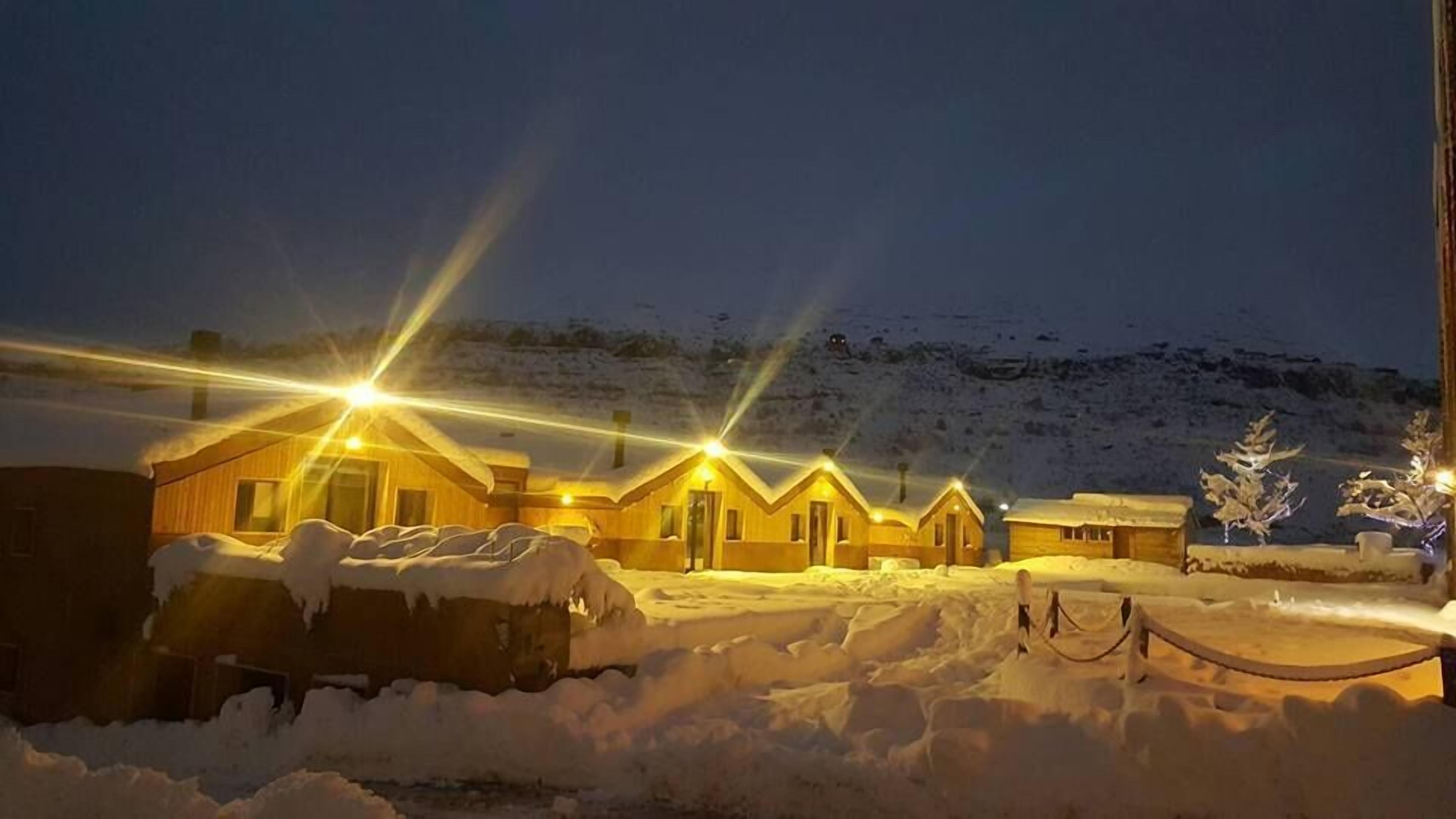 A row of wooden cabins with snow-covered roofs, illuminated by warm lights, set against a snowy landscape.