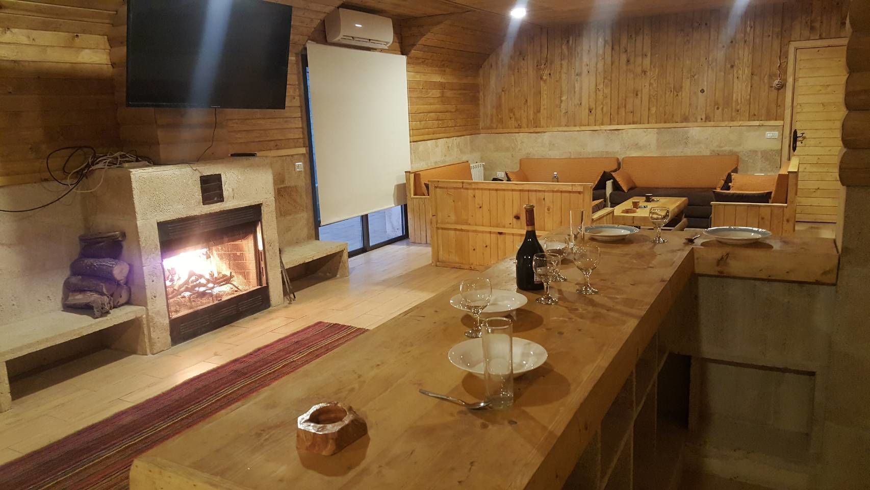 Ein Holzinnenraum mit Kamin, einem langen Esstisch für sechs Personen und einem an der Wand befestigten Fernseher.