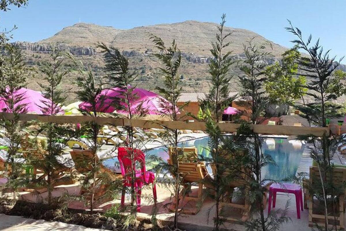 Zona de piscina con sillas de descanso y sombrillas, rodeada de follaje y montañas al fondo.
