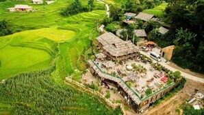An aerial view of a rural village with terraced rice fields, traditional thatched-roof houses, and a central communal area with seating and a small structure. - La Dao Spa Homestay (Ta Van)