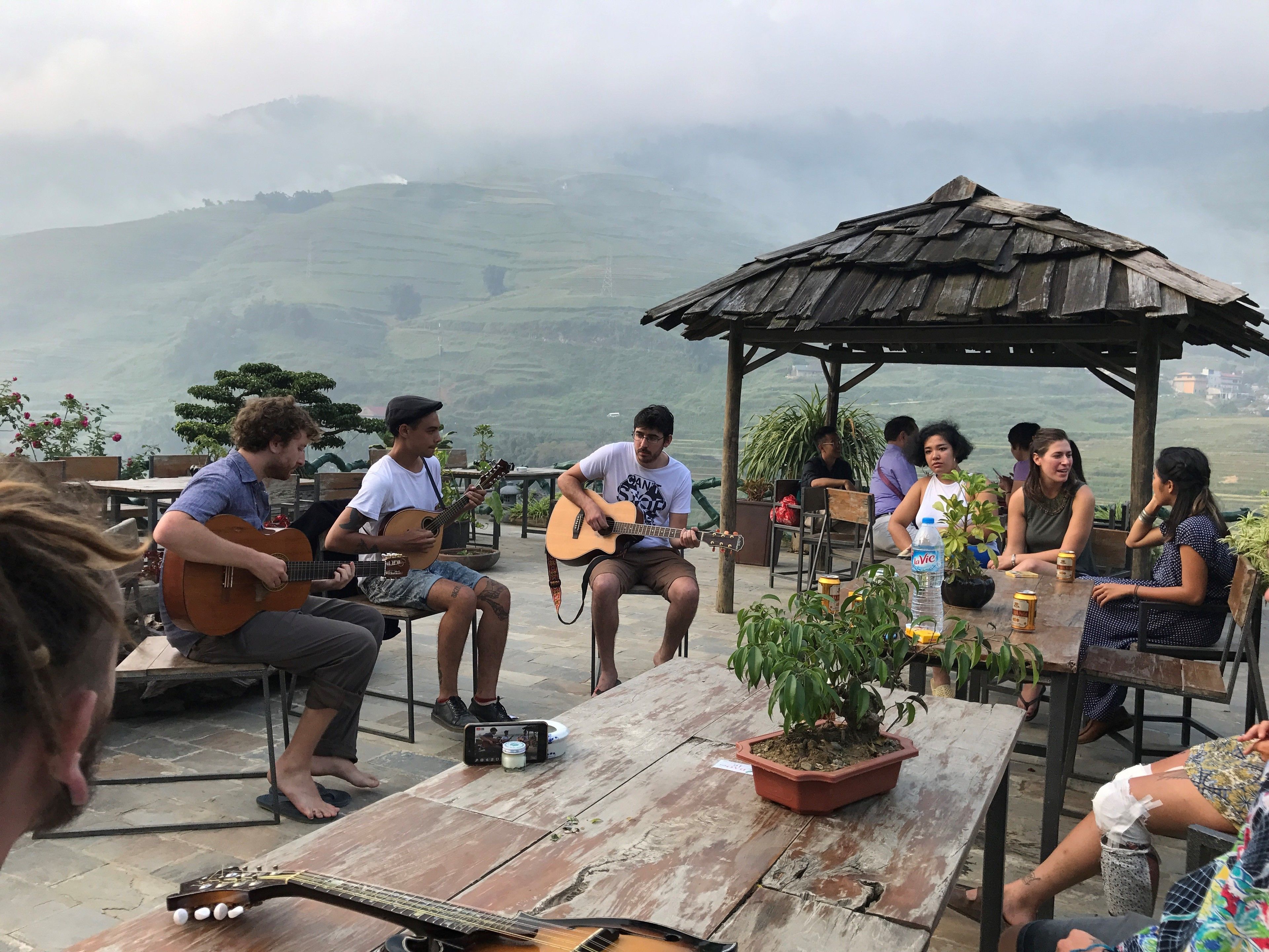 A group of people sitting around a wooden table outdoors, with a guitar player performing. There are potted plants on the table and a gazebo in the background.
