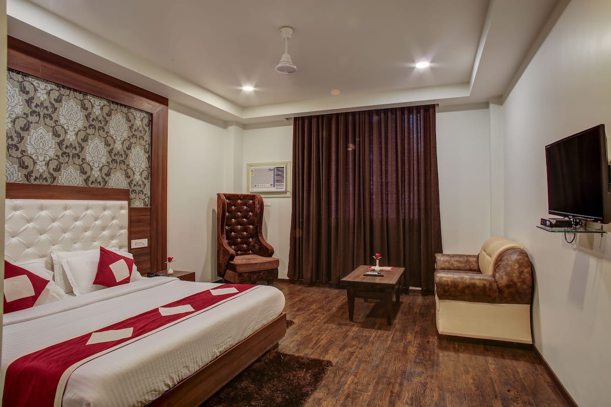 A hotel room with a large bed, a wooden headboard, a brown leather armchair, a beige sofa, a small table with a vase, and a television mounted on the wall.