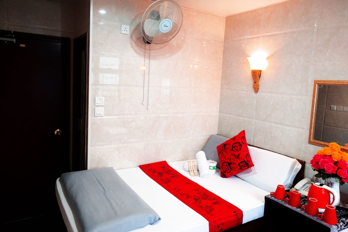 A hotel room with a bed, a bedside table with a phone and red cups, a fan, and a wall-mounted light.
