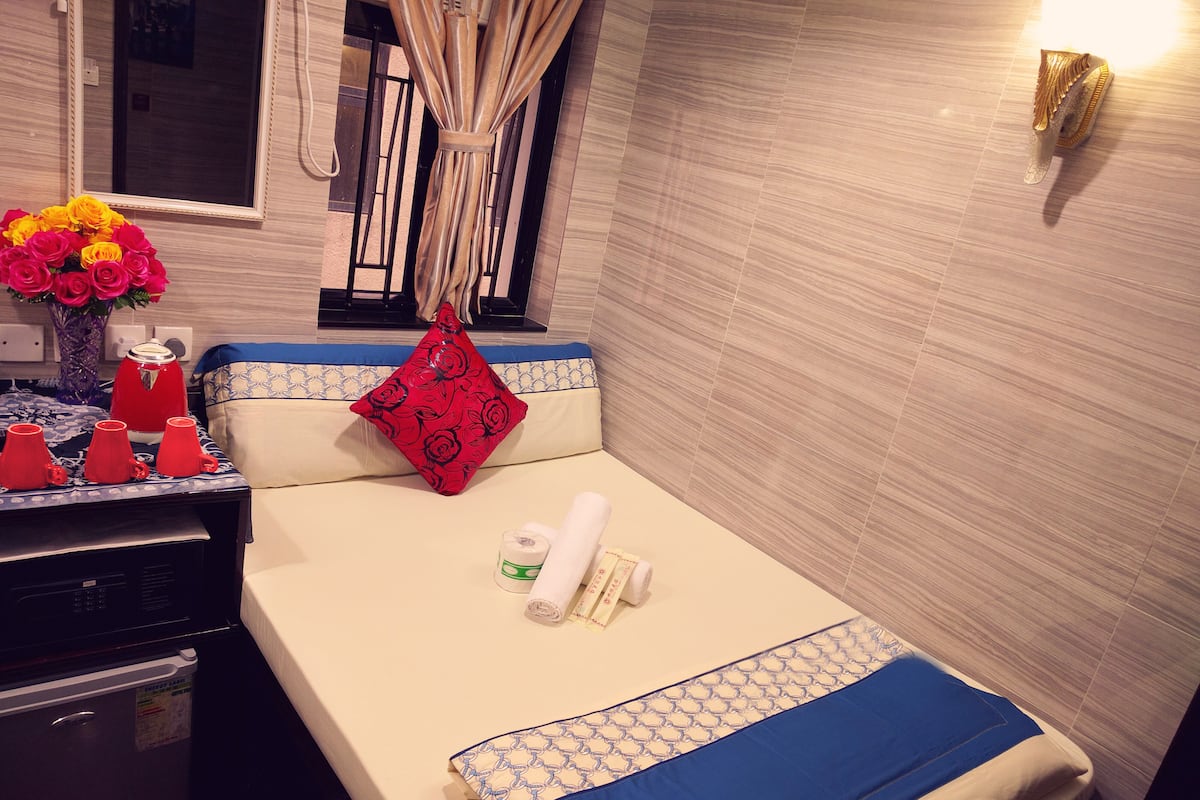 A neatly made bed with a blue and white bedspread, a red pillow with a floral pattern, and a vase of yellow and pink flowers on the nightstand.