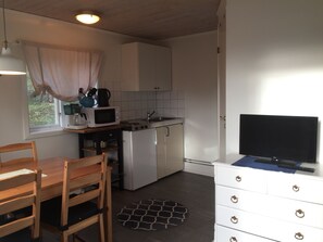 Een kleine keuken met een magnetron, fornuis en spoelbak. Er is een eettafel met stoelen en een witte kast met lades.