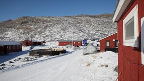 A snowy landscape with red buildings, a clear sky, and a mountain in the background. - Old Camp (Kangerlussuaq)