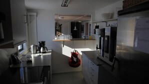 A compact kitchen with a coffee maker, a microwave, and a refrigerator. There is a window providing natural light, and a small shelf with glassware. - Old Camp (Kangerlussuaq)