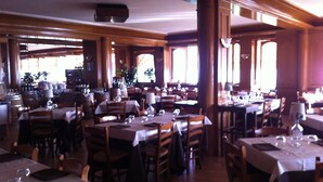 Ein Restaurant mit mehreren Tischen und Stühlen, Holzbalken an der Decke und großen Fenstern, die viel Tageslicht hereinlassen.