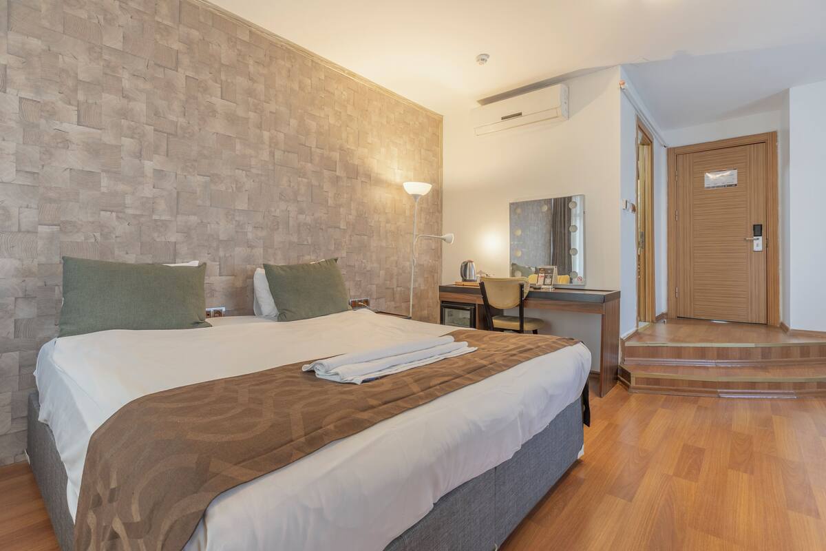A hotel room with a large bed, a desk with a chair, a mirror, and a staircase leading to an entrance.