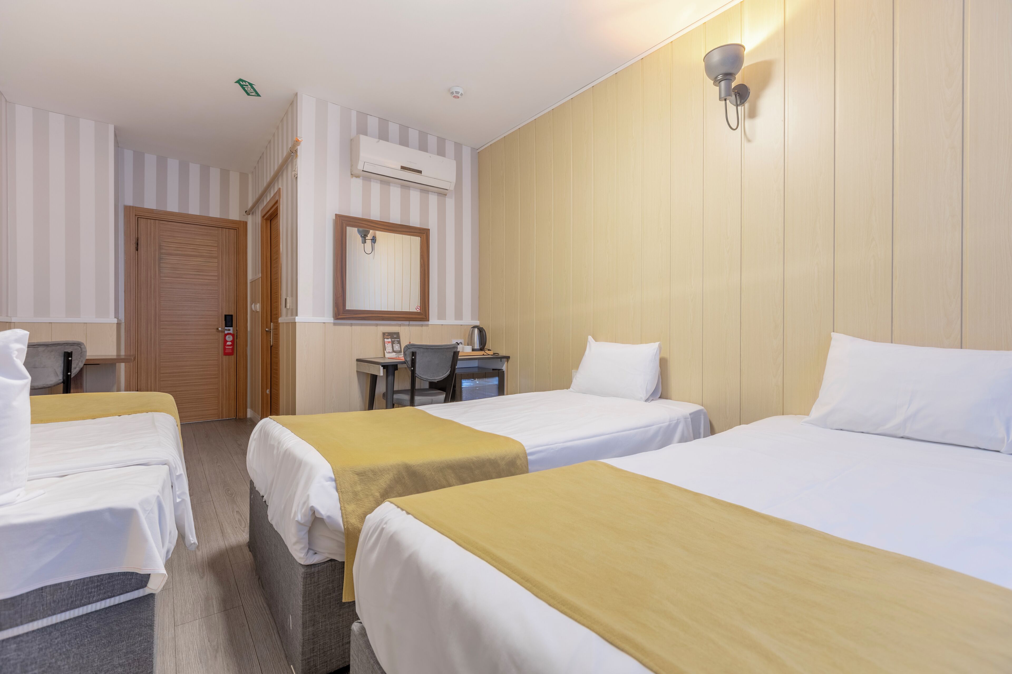 Quarto de hotel com três camas de solteiro, uma mesa de escritório com cadeira, um espelho e ar condicionado.