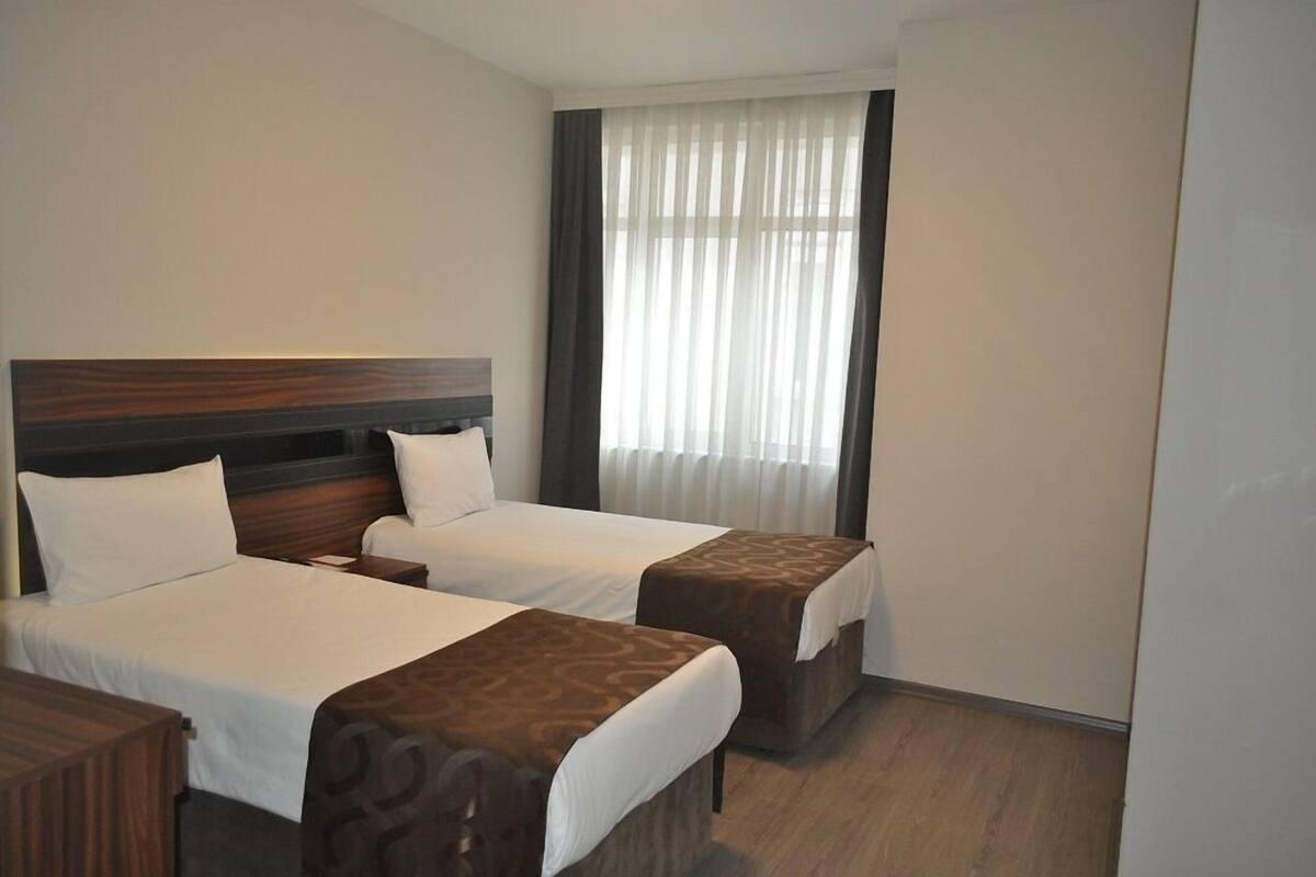 A hotel room with two beds, a wooden headboard, a window with sheer curtains, and a wooden floor.