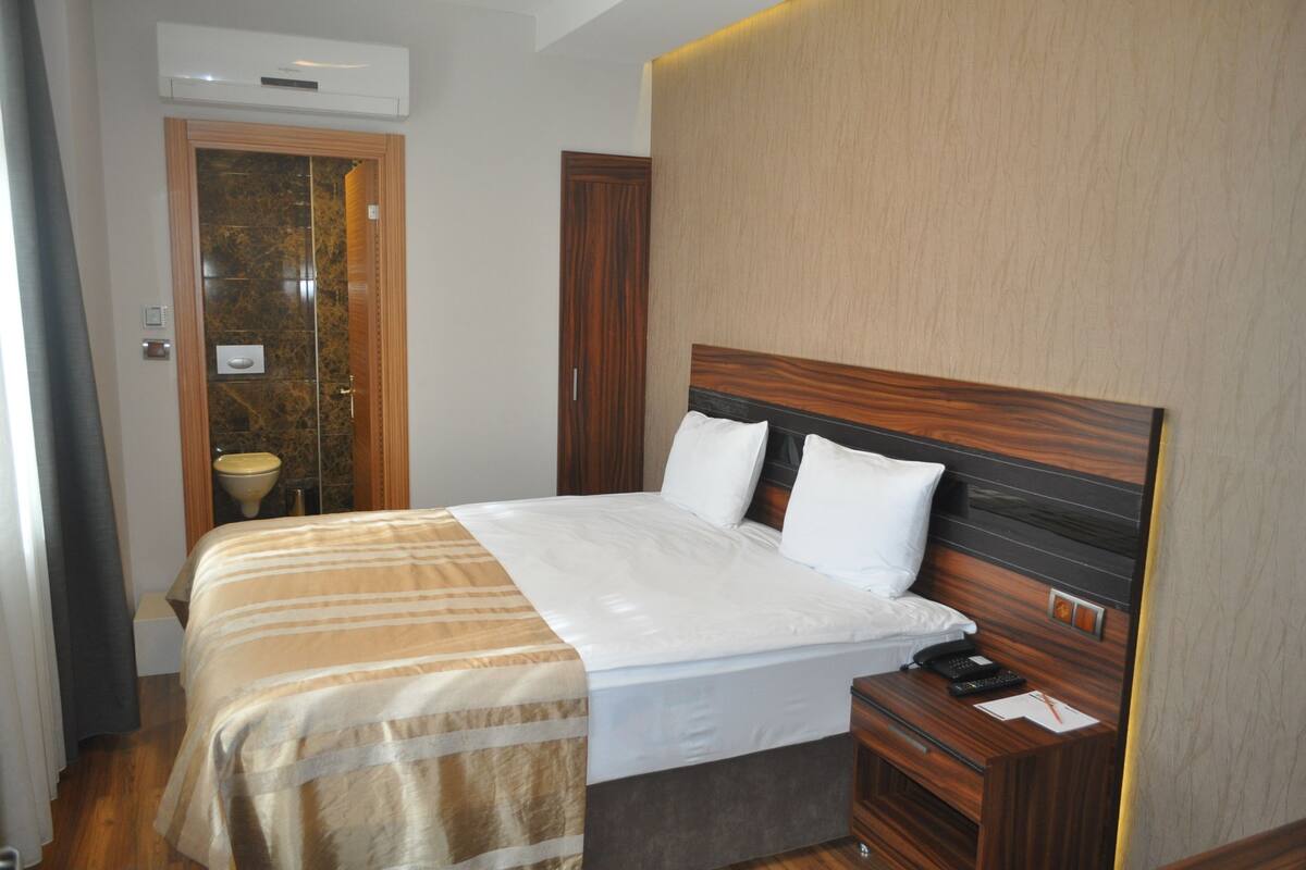 A hotel room with a bed, a bedside table with a phone and a remote, a wall-mounted air conditioner, and a bathroom with a sink and mirror.