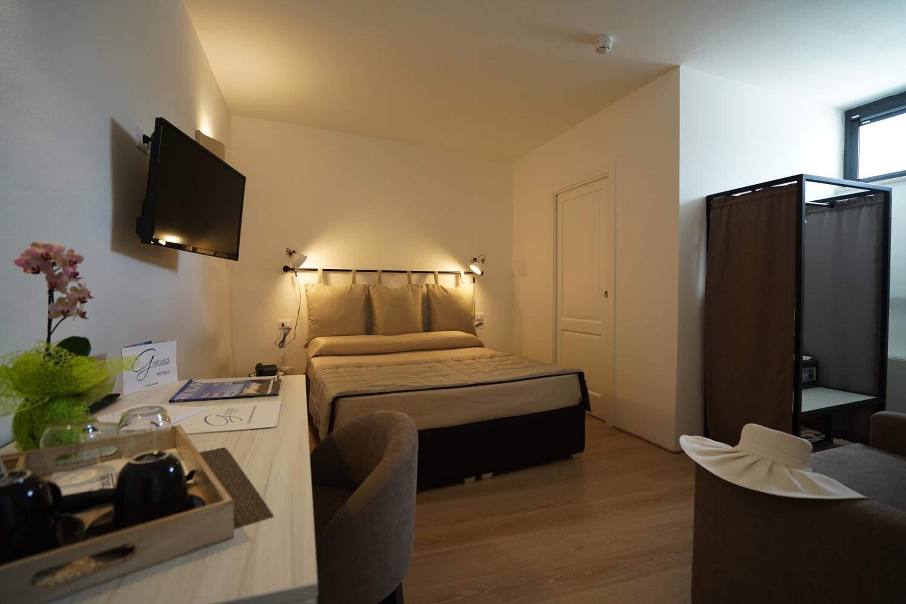Quarto de hotel com cama, mesa de cabeceira, televisão e um pequeno espaço de estar.