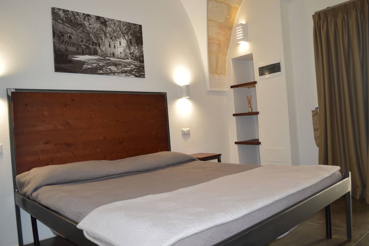 Un lit bien fait, avec une tête de lit en bois, une étagère fixée au mur ornée d’une petite plante, et un tableau encadré au-dessus du lit.