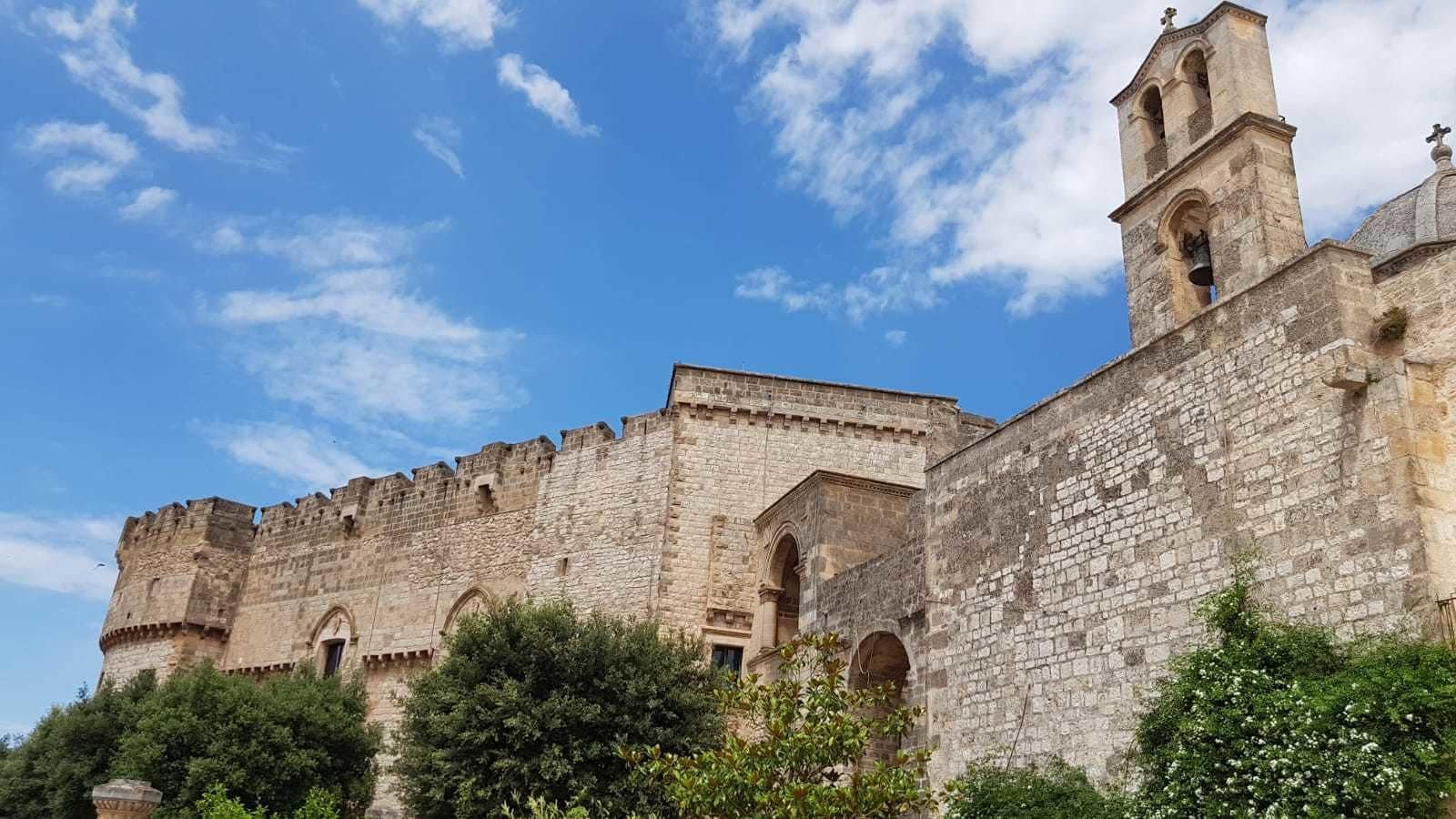 Un edificio histórico de piedra con una torre de campanas, ventanas de arco y una muralla fortificada.