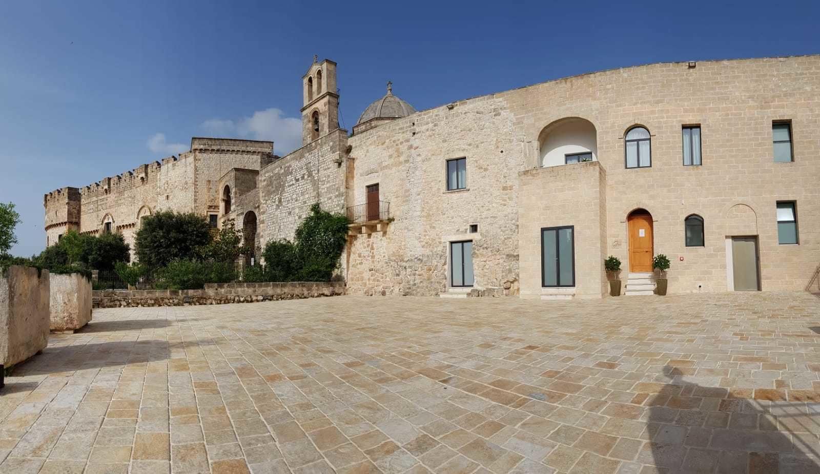 Un patio con pavimento de piedra, un edificio histórico con una torre de campanas y una entrada moderna con puerta de madera.
