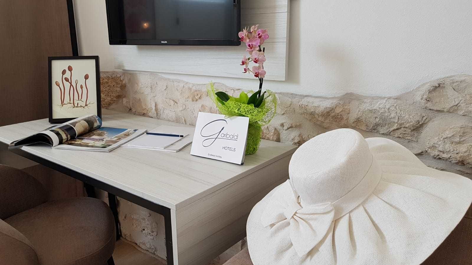 A hotel desk with a framed picture, a magazine, a notepad, a pen, a vase with green foliage, a pink orchid, and a sign reading "Gianluca Hotels."