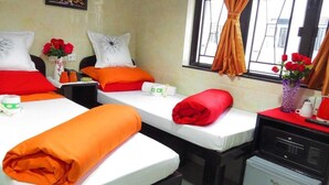 Une chambre d’hôtel avec deux lits, chacun agrémenté de draps rouges et oranges, une petite table sur laquelle est posé un vase de fleurs rouges, un four à micro-ondes et une fenêtre avec des rideaux.