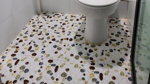Une salle de bain avec des toilettes blanches, un lavabo et une douche. Le sol est recouvert de carreaux imitant des galets.