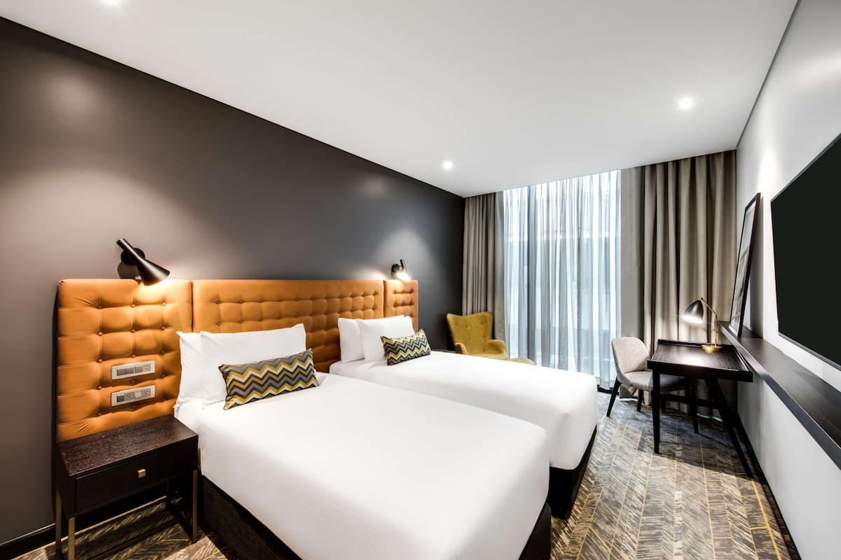 Ein Hotelzimmer mit zwei Betten, einem Schreibtisch, einem Stuhl und einem großen Fenster mit Vorhängen.