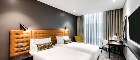 Ein Hotelzimmer mit zwei Betten, einem Schreibtisch, einem Stuhl und einem großen Fenster mit Vorhängen.