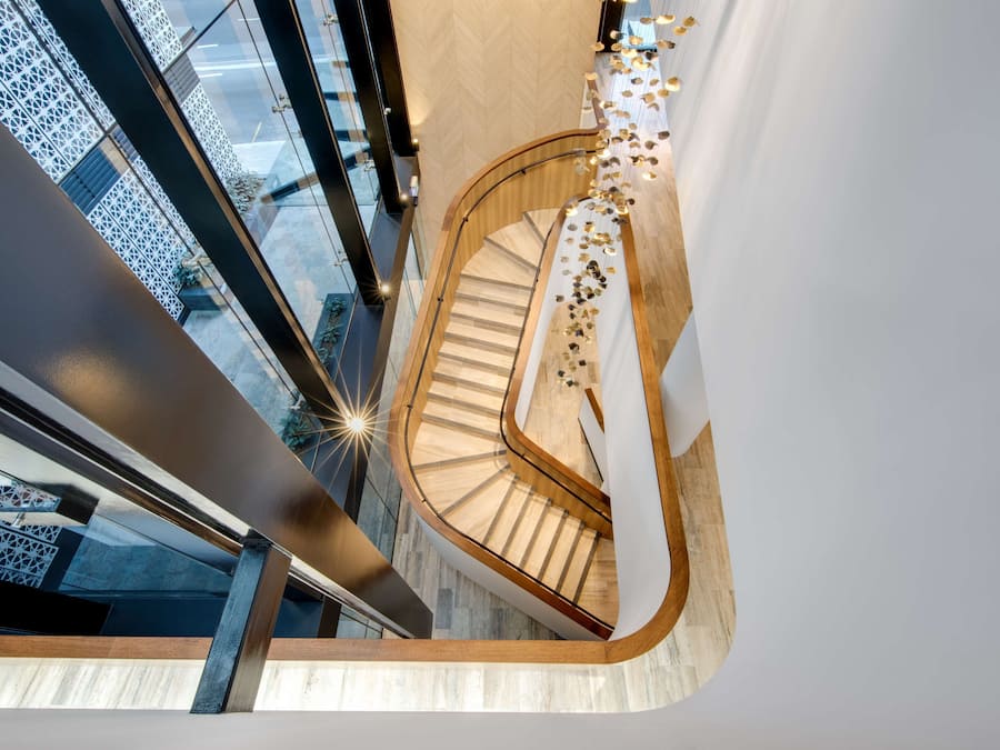 A modern spiral staircase with a wooden finish and glass railings, leading to an upper floor with a view of the cityscape.