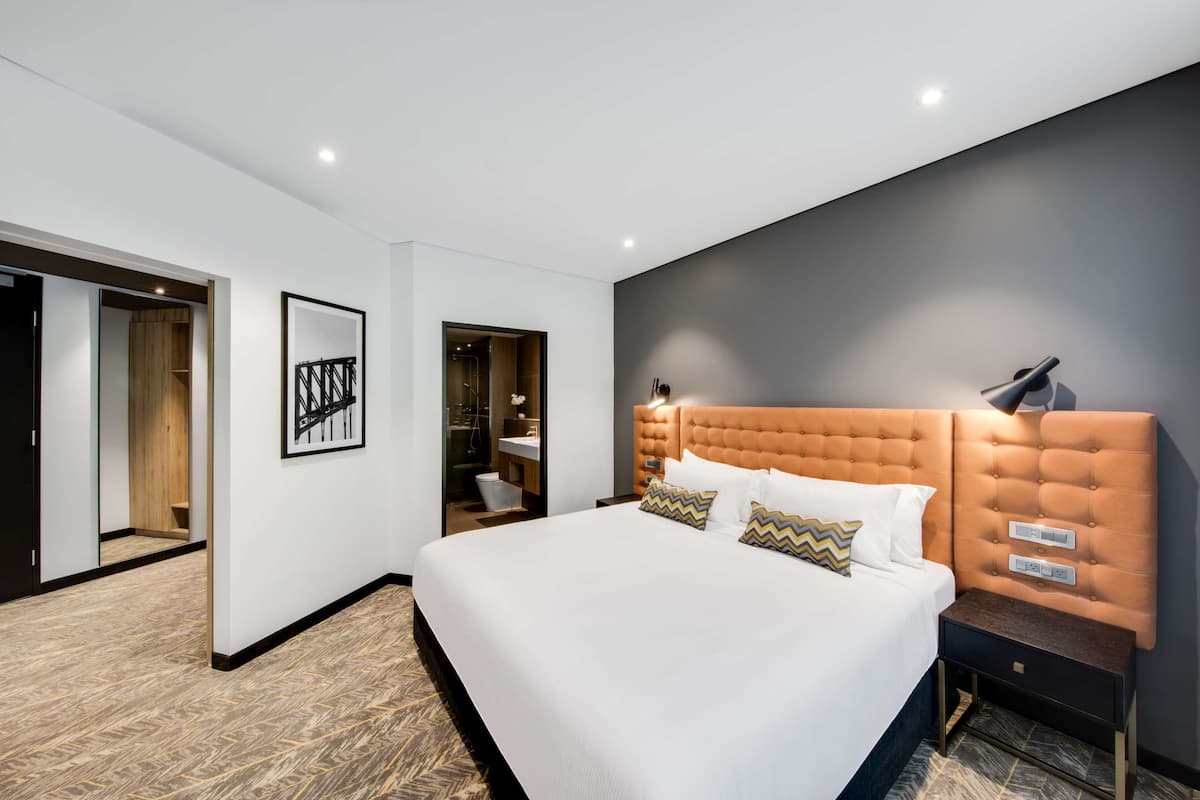Ein modernes Hotelzimmer mit einem großen Bett, einem Kopfteil mit integrierten Bedienelementen, einem Nachttisch und einem gerahmten Kunstwerk an der Wand.