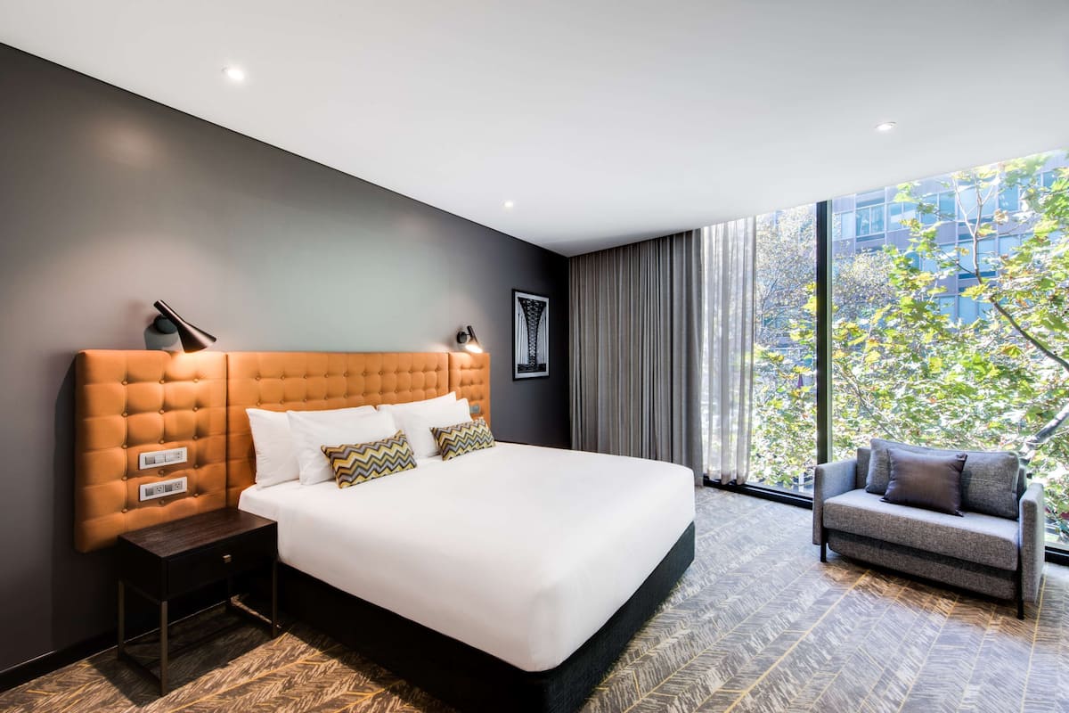 Ein modernes Hotelzimmer mit einem großen Bett, einem Nachttisch, einer Sitzecke und einem großen Fenster mit Blick ins Freie.