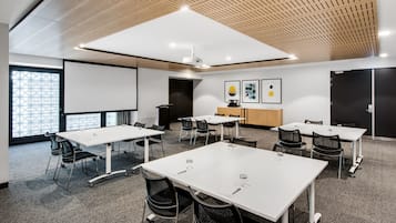 Una sala de conferencias moderna con mesas y sillas blancas, una pantalla de proyector y techo de madera.