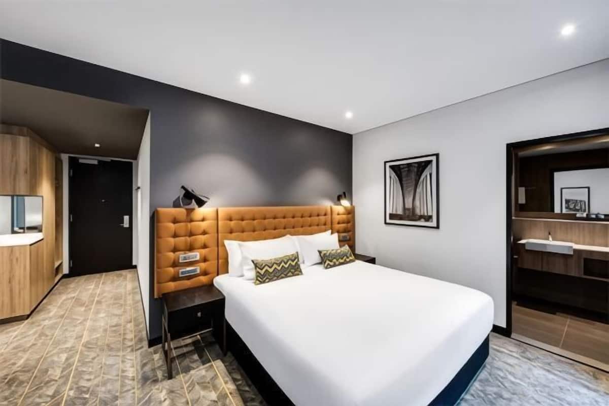 Ein modernes Hotelzimmer mit einem großen Bett, einem Holzschrank, einem Badezimmer mit Spiegel und Waschbecken sowie einem gerahmten Bild an der Wand.