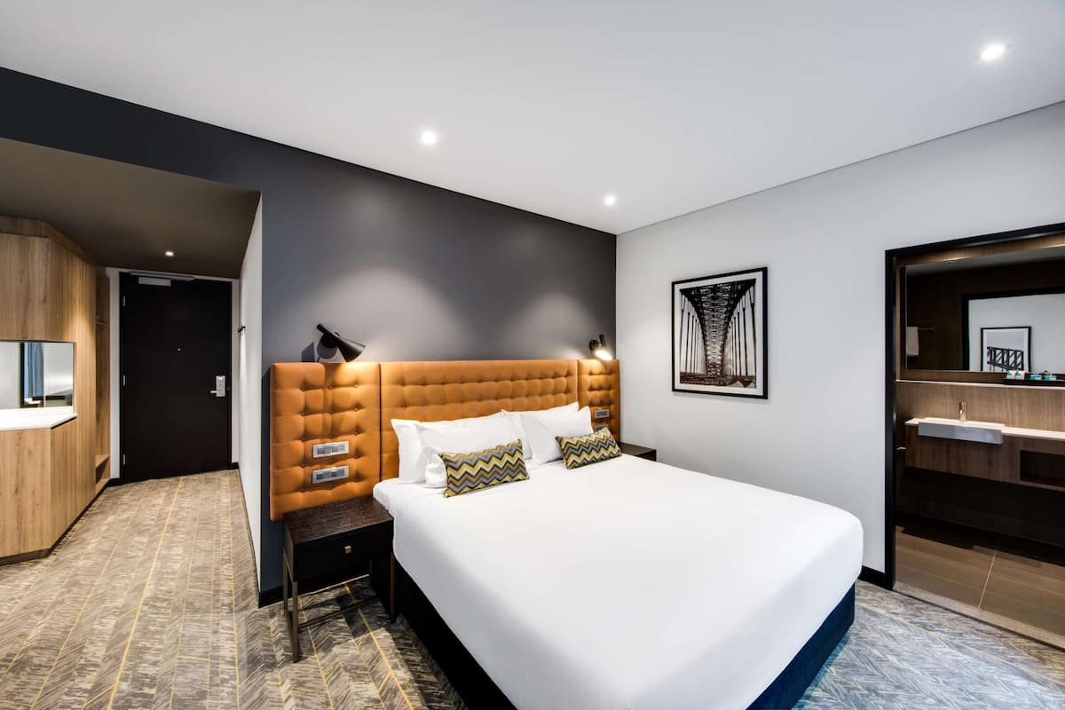 Ein modernes Hotelzimmer mit einem großen Bett, einem Holzschrank, einem Badezimmer mit Spiegel und einem Nachttisch mit Lampe.