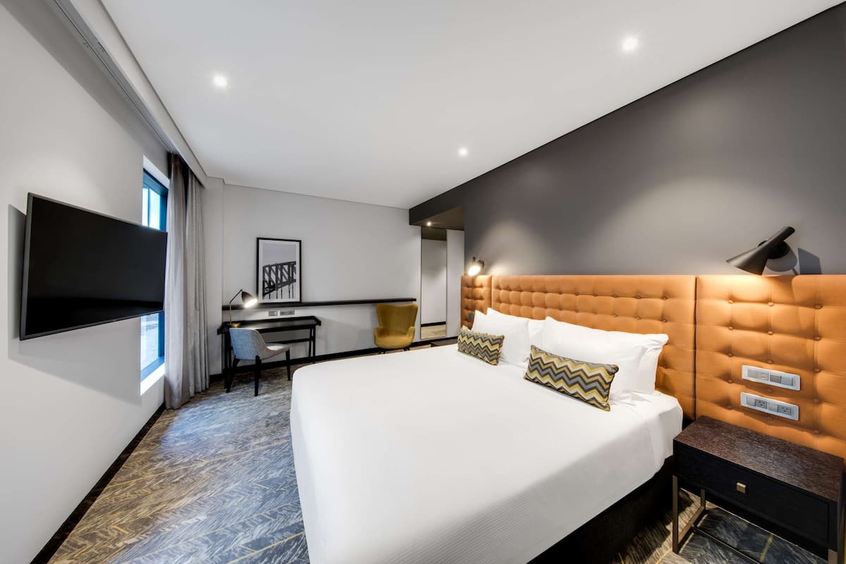 Ein modernes Hotelzimmer mit einem großen Bett, einem Schreibtisch, einem Sessel und einem an der Wand montierten Fernseher.