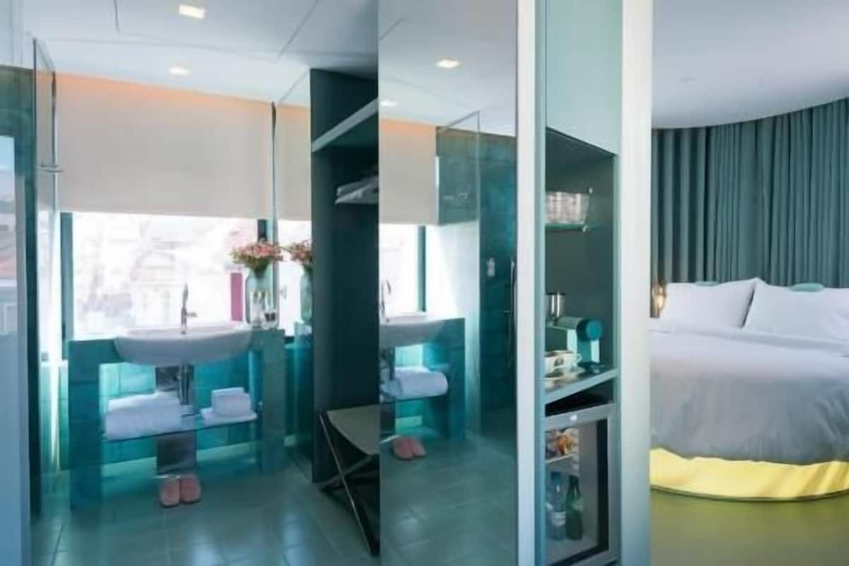 Una habitación de hotel moderna con cama, baño con paredes de vidrio que incluye un lavamanos y un espejo, y vistas a la ciudad.