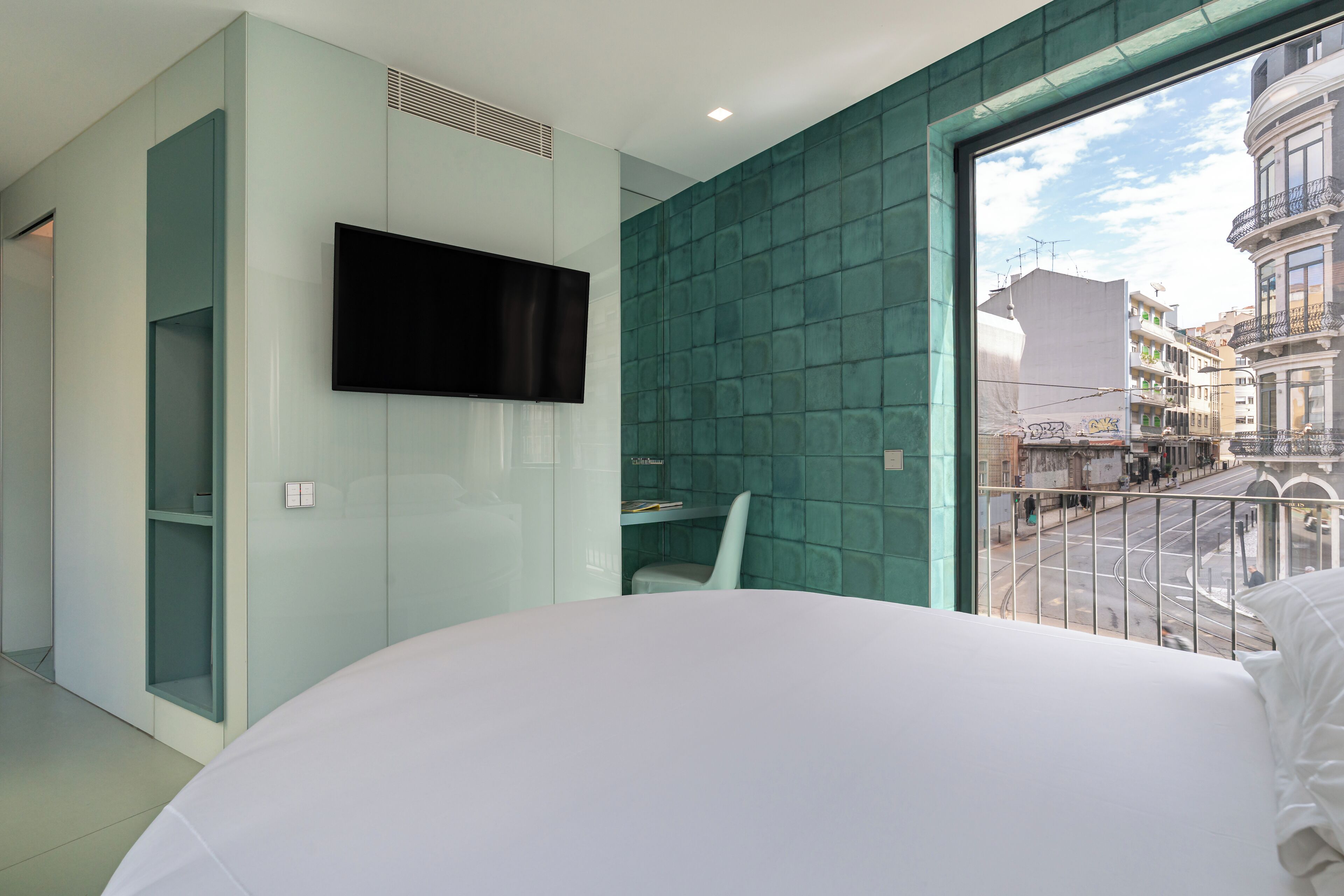 Una habitación de hotel moderna con una cama grande, un televisor montado en la pared, un escritorio pequeño y vistas de la ciudad a través de un ventanal amplio.