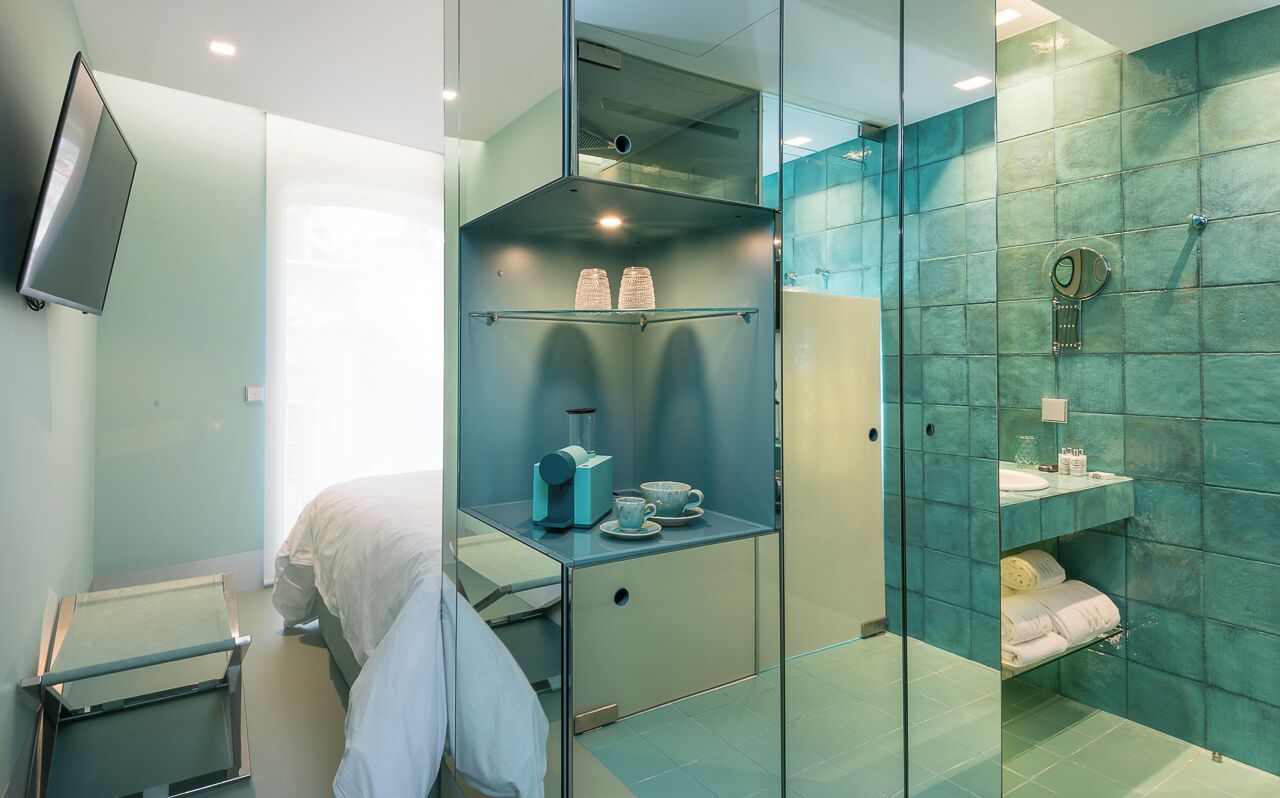 Una habitación de hotel moderna con cama, tocador con paredes de vidrio y zona de ducha con pared de azulejos verdes.
