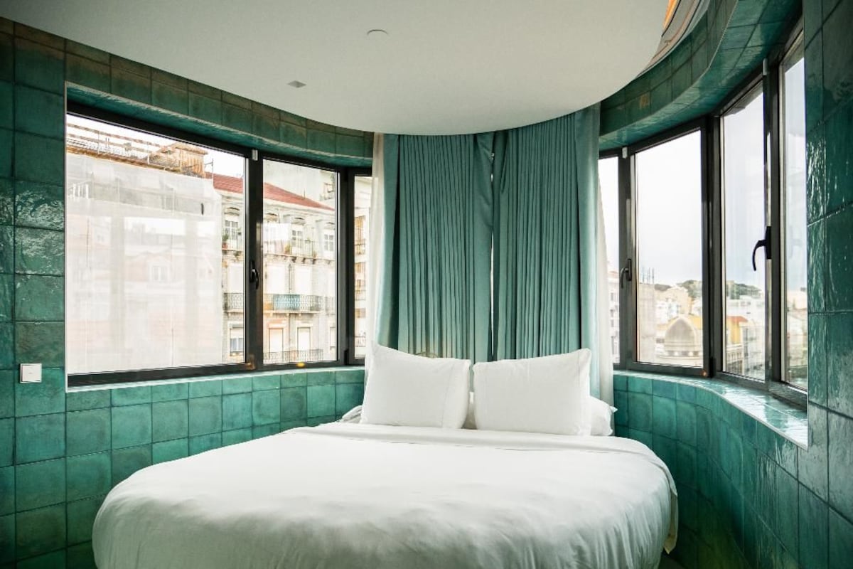 Une chambre d’hôtel moderne avec un lit aux lignes courbes, de grandes fenêtres et des murs de couleur turquoise.