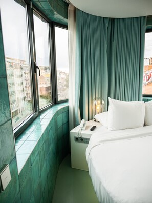 A hotel room with a bed, a bedside table with a phone and remote, a window with curtains, and a view of buildings outside. - WC by The Beautique Hotels (Lisbon)