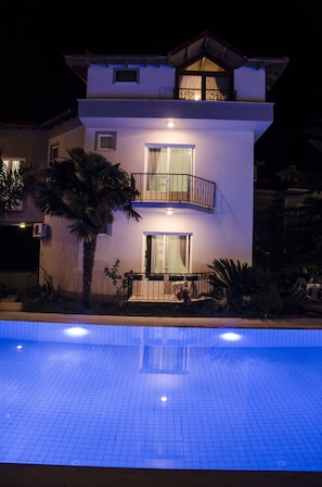 A modern two-story house with a balcony, a swimming pool in the foreground, and a well-lit exterior.