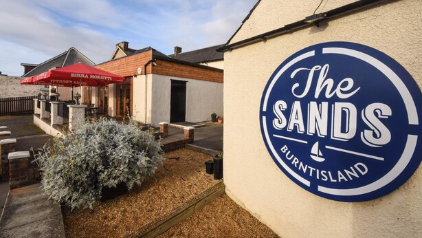 Un edificio con un cartel que dice "The Sands Burntisland" y un paraguas rojo con la leyenda "Birra Moretti".