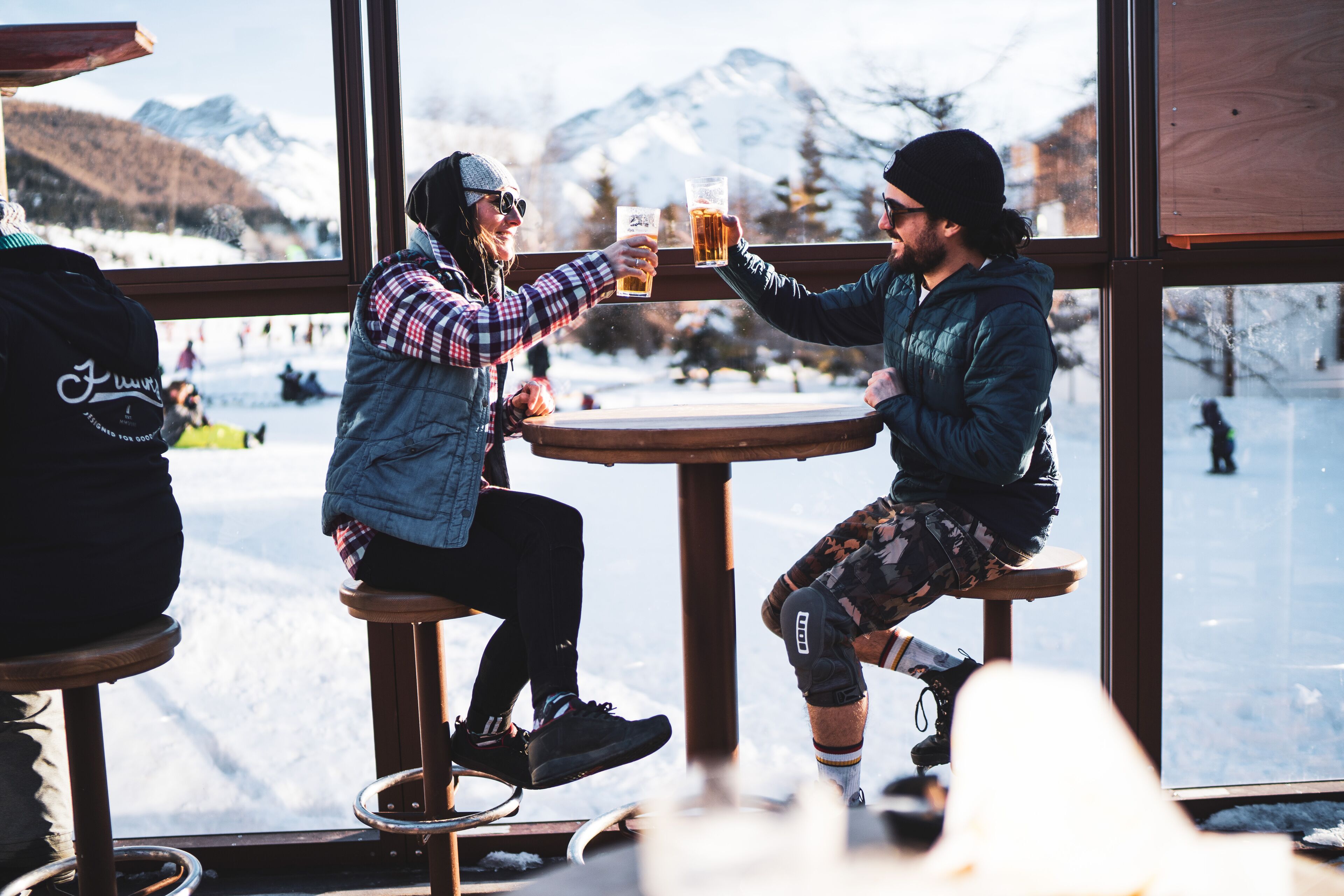 Two people toasting with beer glasses at a wooden table with a view of snow-covered mountains through a window.